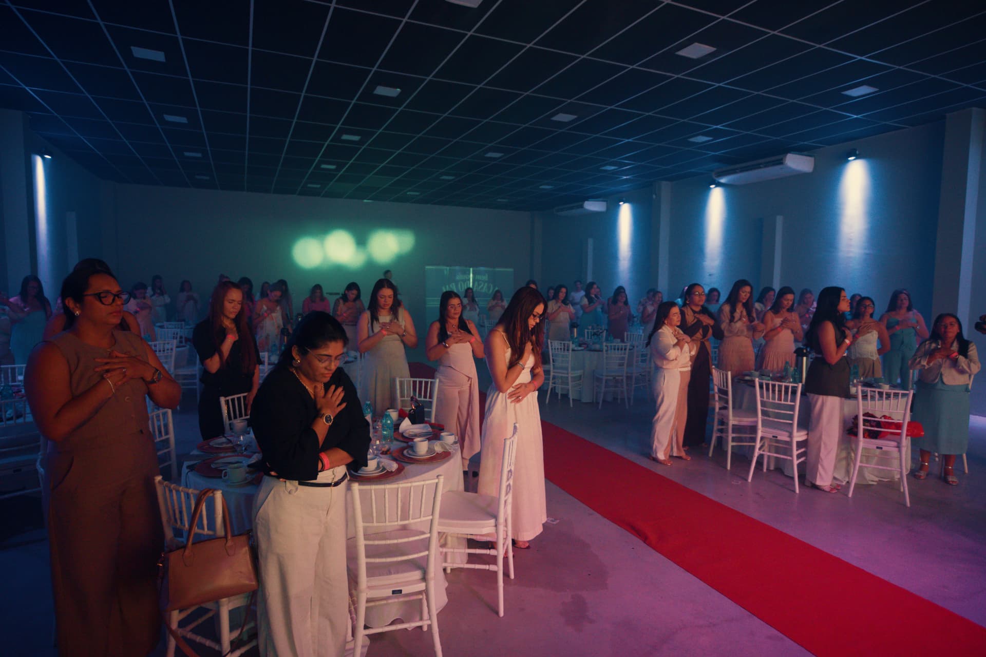 Salão do encontro com mulheres de pé, em oração, com as mãos sobre o peito