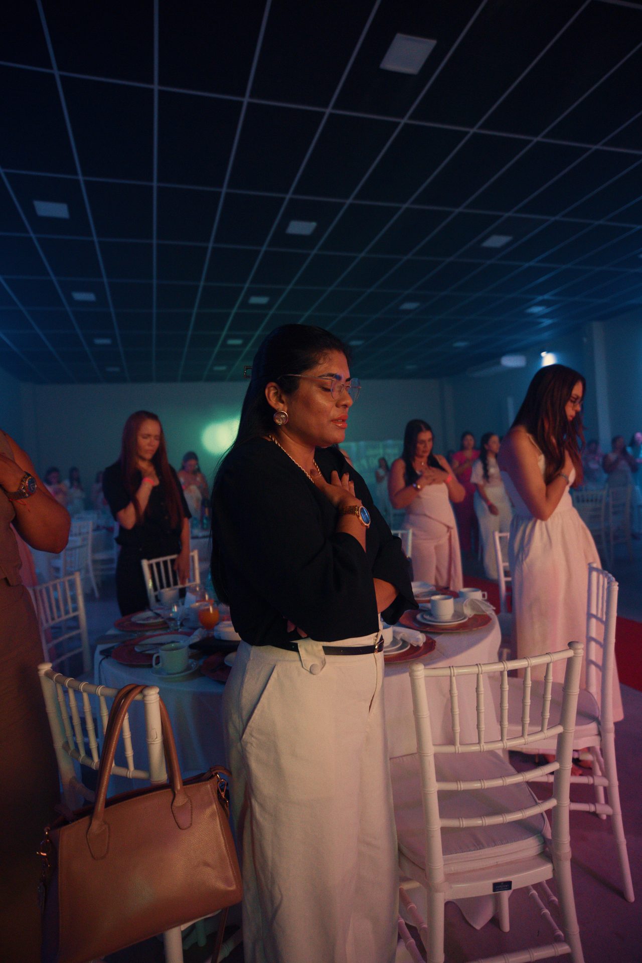 Participantes de pé entre as mesas, de olhos fechados e mãos sobre o coração